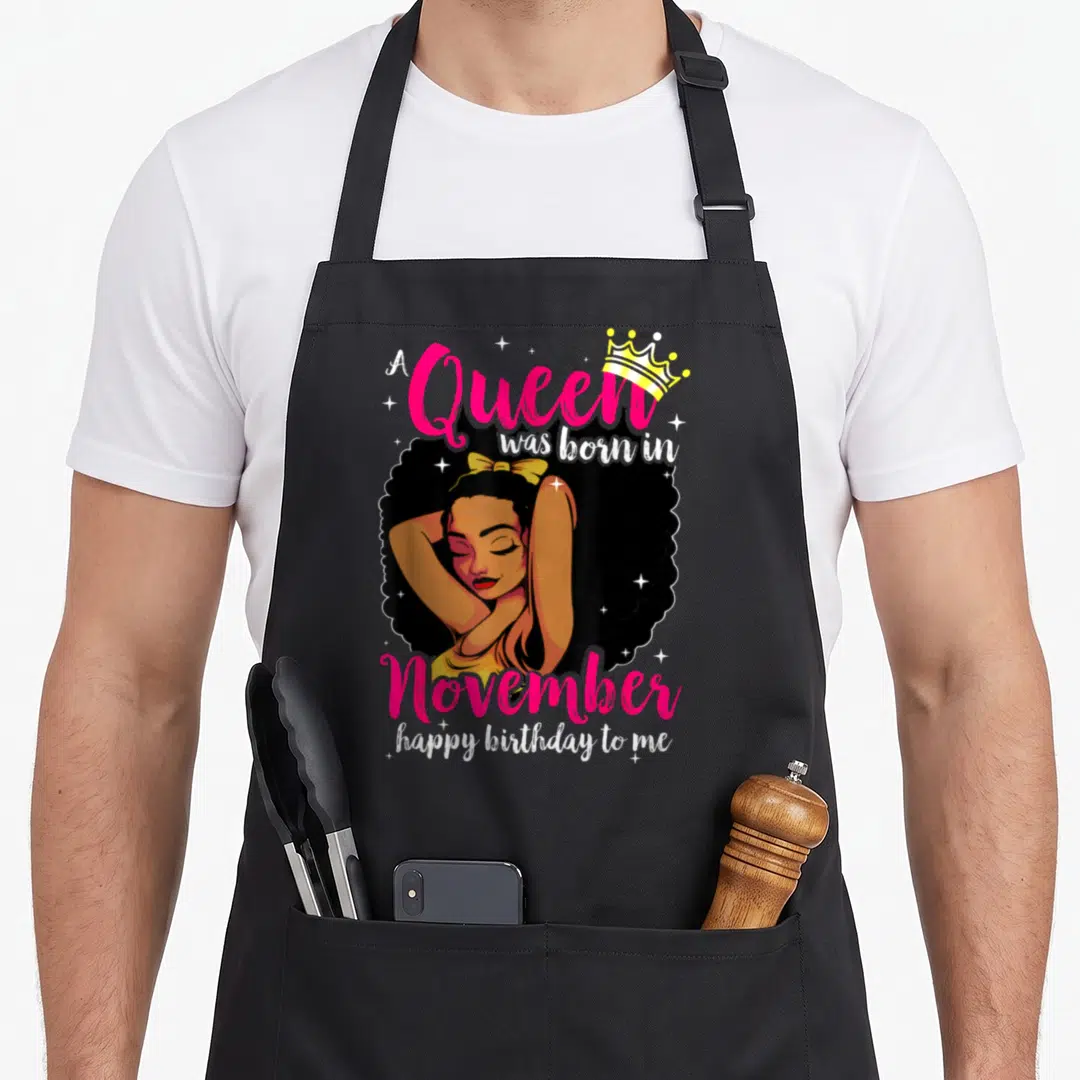 Afro diva queen birthday apron - Cartoon woman with crown bow and A Queen was born in November happy birthday to me text — shown on a black cotton canvas apron against a white background