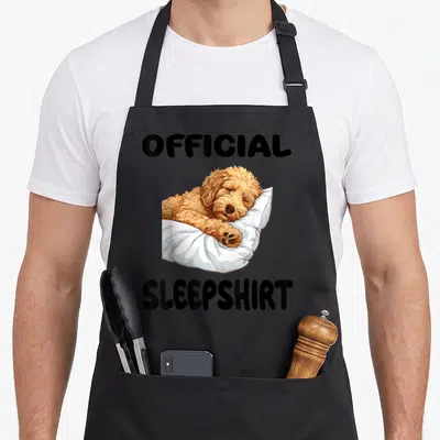 Goldendoodle sleepshirt dog apron - Cartoon tan dog sleeping on white pillow with black OFFICIAL SLEEPSHIRT text — shown on a black cotton canvas apron against a white background