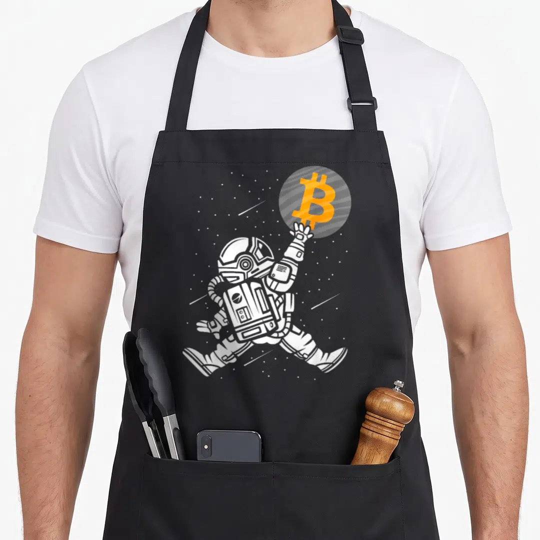 Astronaut bitcoin apron - White cartoon astronaut reaching for orange Bitcoin symbol inside grey planet with stars — shown on a black cotton canvas apron against a white background