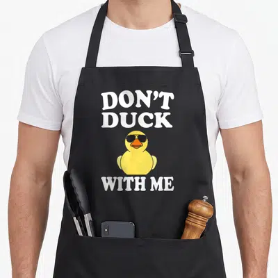 Funny rubber duck apron - Yellow cartoon duck with black sunglasses and white text Don't Duck With Me — shown on a black cotton canvas apron against a white background