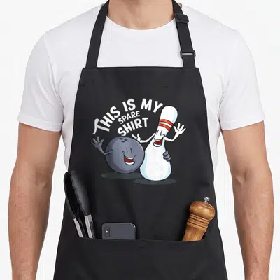 Spare game aprons - Cartoon ball hugging standing pin with arched text THIS IS MY SPARE SHIRT — shown on a black cotton canvas apron against a white background
