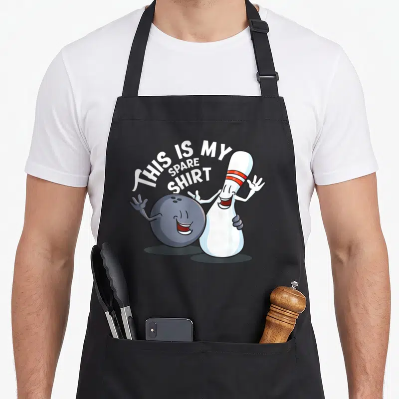 Spare game aprons - Cartoon ball hugging standing pin with arched text THIS IS MY SPARE SHIRT — shown on a black cotton canvas apron against a white background