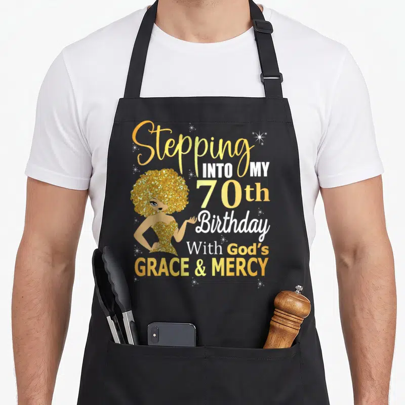 70th grace apron - Cartoon woman with gold glitter hair next to stacked text about 70th birthday and God's grace — shown on a black cotton canvas apron against a white background