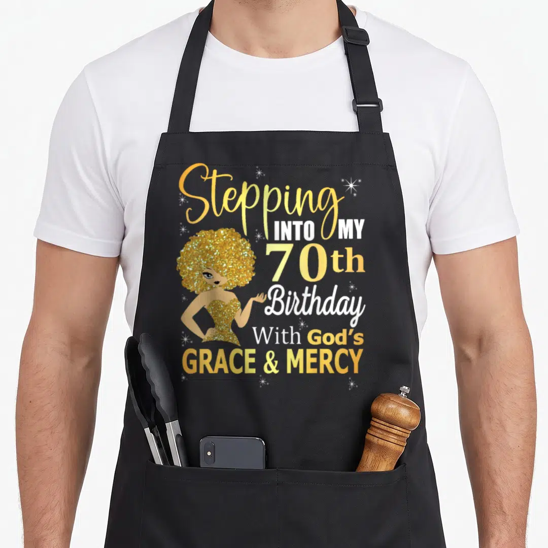 70th grace apron - Cartoon woman with gold glitter hair next to stacked text about 70th birthday and God's grace — shown on a black cotton canvas apron against a white background