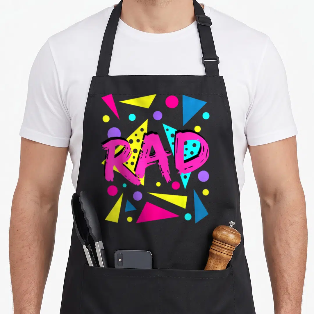 Rad eighty 1980s apron - Pink RAD text over yellow blue and purple geometric triangles and circles — shown on a black cotton canvas apron against a white background