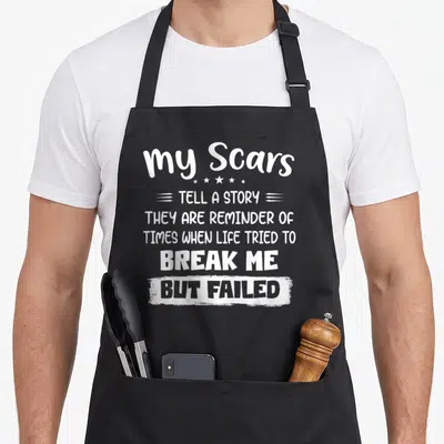 Scar the story of the apron - White typography reading my Scars tell a story with star icons and brushstroke box — shown on a black cotton canvas apron against a white background