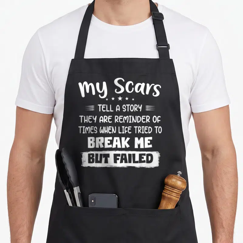Scar the story of the apron - White typography reading my Scars tell a story with star icons and brushstroke box — shown on a black cotton canvas apron against a white background