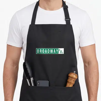 Street theater apron - Green street sign reading BROADWAY next to a Statue of Liberty outline — shown on a black cotton canvas apron against a white background