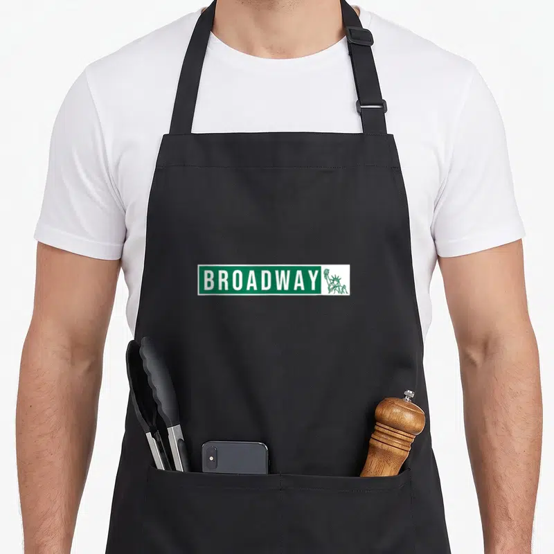 Street theater apron - Green street sign reading BROADWAY next to a Statue of Liberty outline — shown on a black cotton canvas apron against a white background