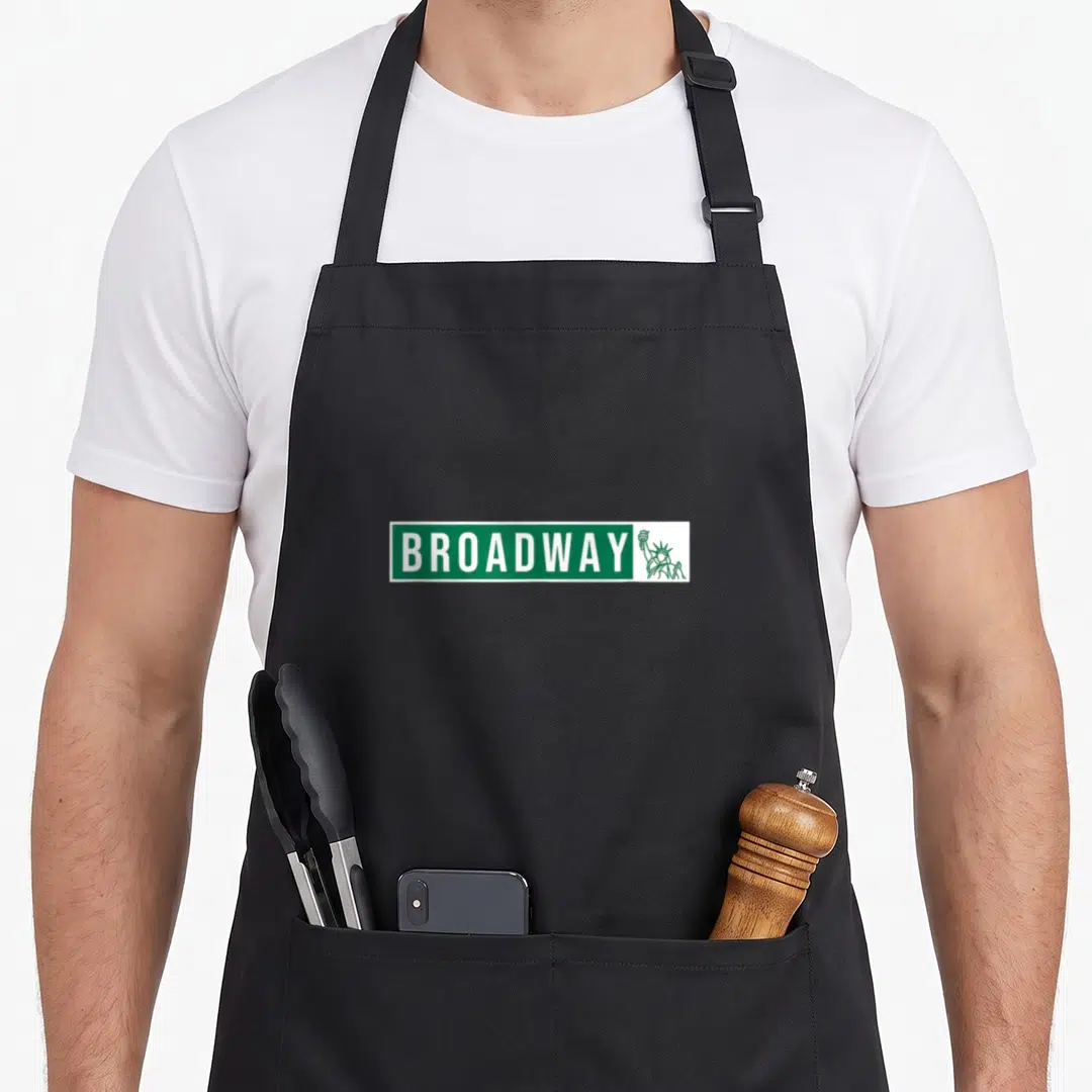 Street theater apron - Green street sign reading BROADWAY next to a Statue of Liberty outline — shown on a black cotton canvas apron against a white background