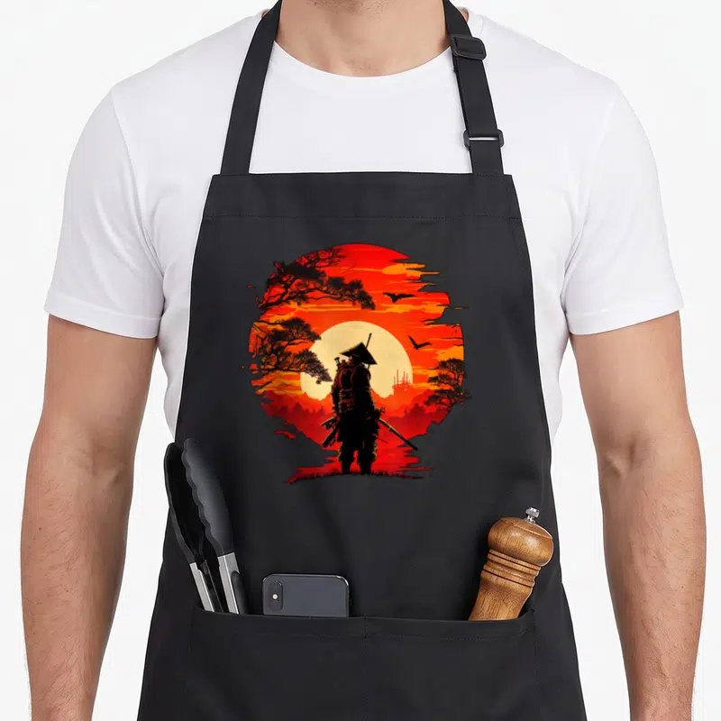Japanese martial apron - Black samurai silhouette standing before a yellow sun inside a red circle with black trees — shown on a black cotton canvas apron against a white background