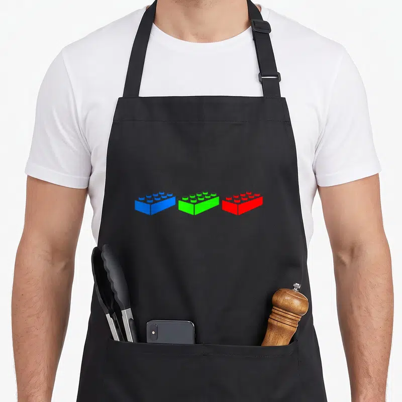 Construction block & apron - Three toy bricks in blue, green, and red arranged horizontally in a minimalist style — shown on a black cotton canvas apron against a white background
