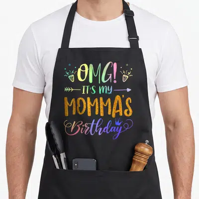 Mommas tie-dye apron - Stacked birthday text with gold glitter momma, rainbow gradient words, party poppers, arrow — shown on a black cotton canvas apron against a white background
