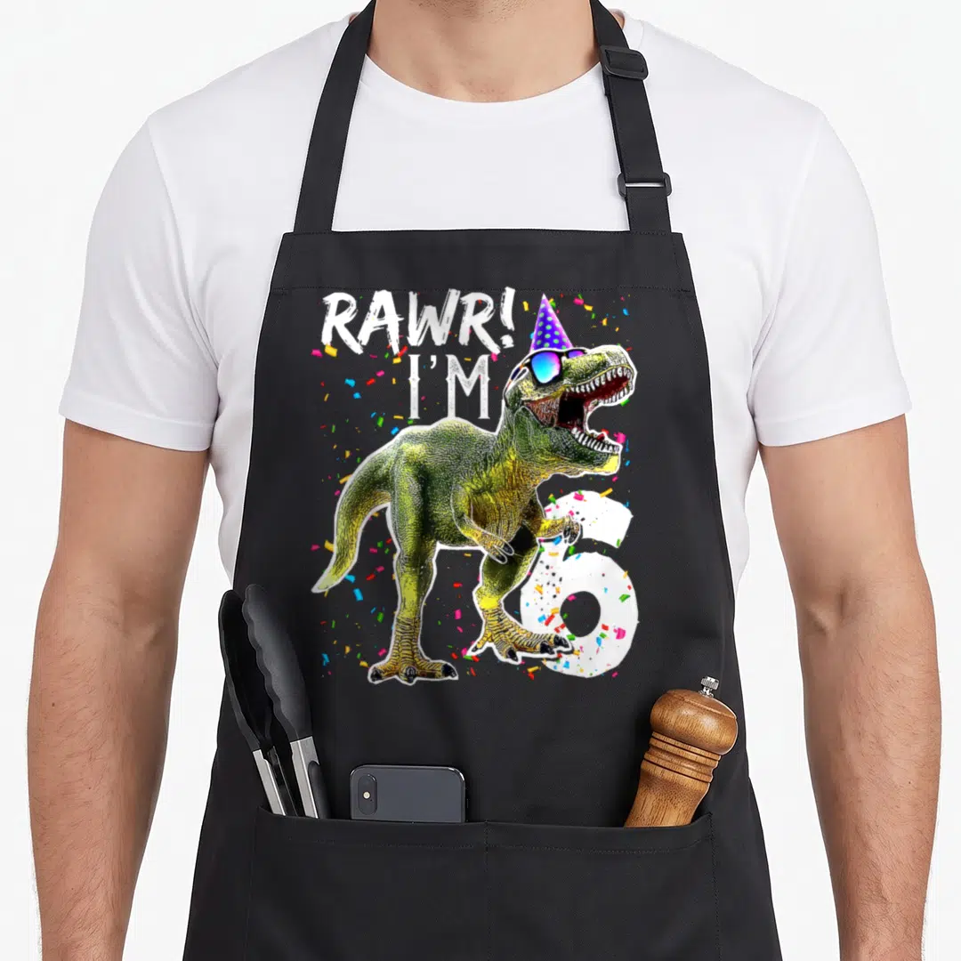 6th t-rex apron - Cartoon T-Rex in party hat and sunglasses with RAWR! I'M 6 text and confetti — shown on a black cotton canvas apron against a white background