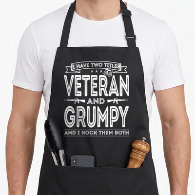 Grumpy father day apron - White stacked text reading I Have Two Titles Veteran And Grumpy with rifles and dog tags — shown on a black cotton canvas apron against a white background