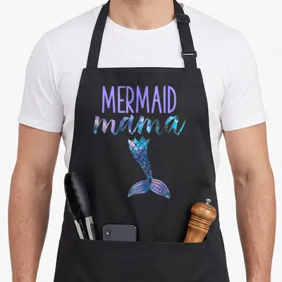 Mermaid birthday mama apron - MERMAID mama text above purple blue pink glittery mermaid tail on black bib — shown on a black cotton canvas apron against a white background