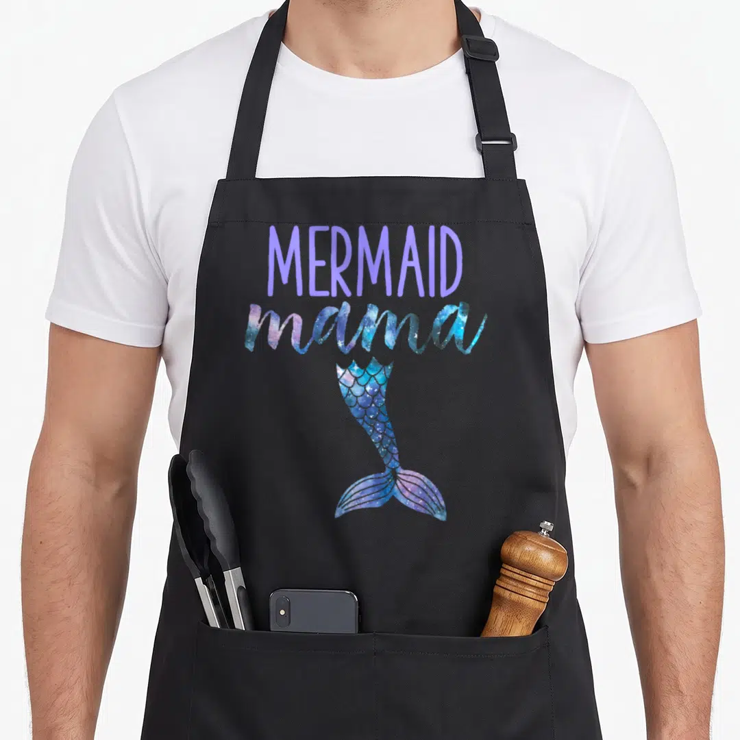 Mermaid birthday mama apron - MERMAID mama text above purple blue pink glittery mermaid tail on black bib — shown on a black cotton canvas apron against a white background