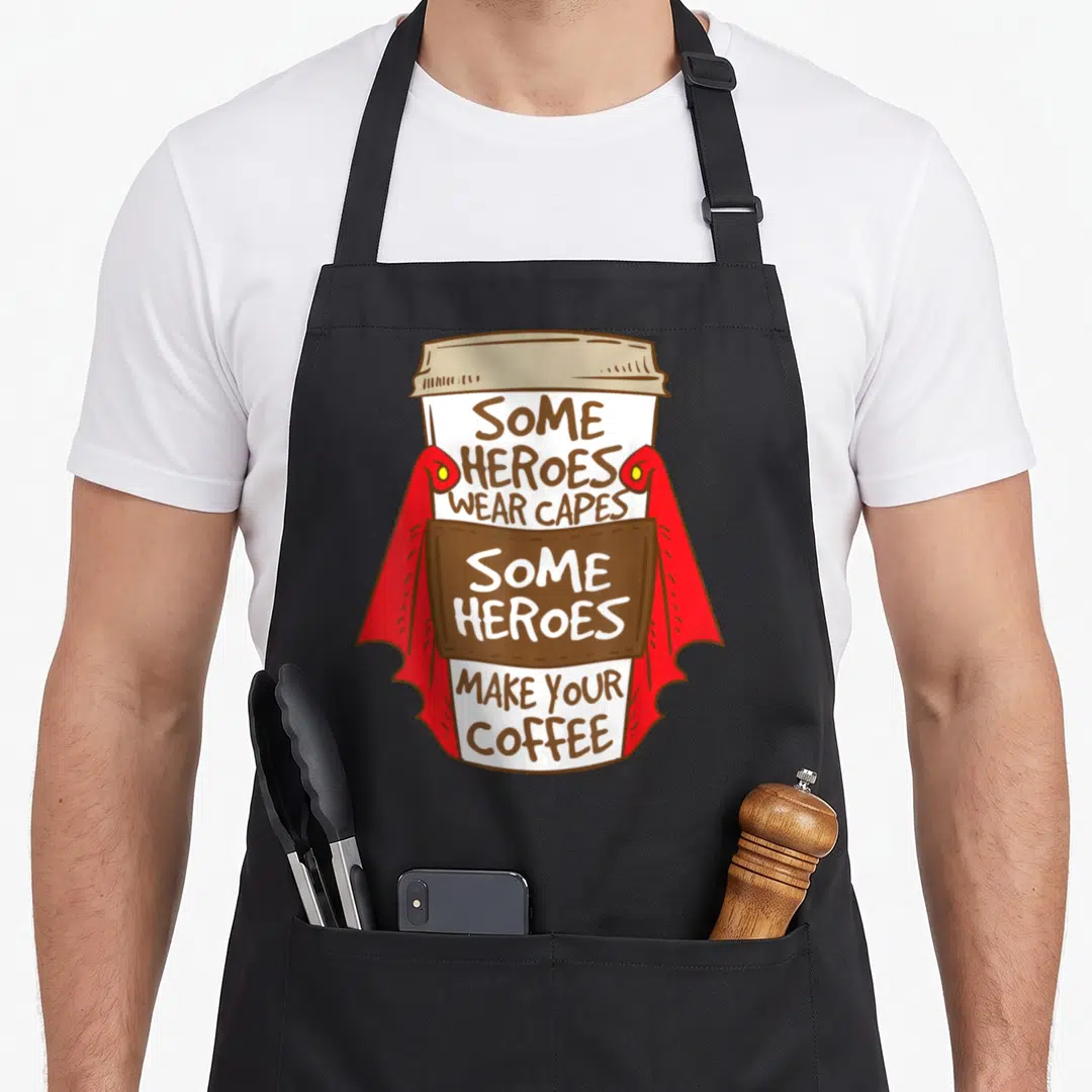 Superhero coffee apron - Cartoon cup with red cape and text SOME HEROES WEAR CAPES SOME HEROES MAKE YOUR COFFEE — shown on a black cotton canvas apron against a white background