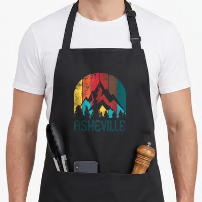 Vintage asheville apron - Grey mountain silhouette against yellow orange red and teal striped sun with teal trees — shown on a black cotton canvas apron against a white background
