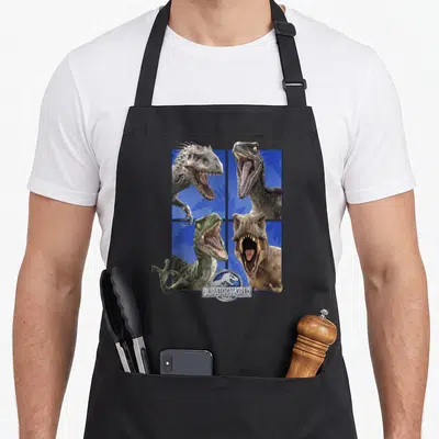 Roar dinosaur apron - Four dinosaur heads in a grid over blue background with JURASSIC WORLD logo at bottom center — shown on a black cotton canvas apron against a white background