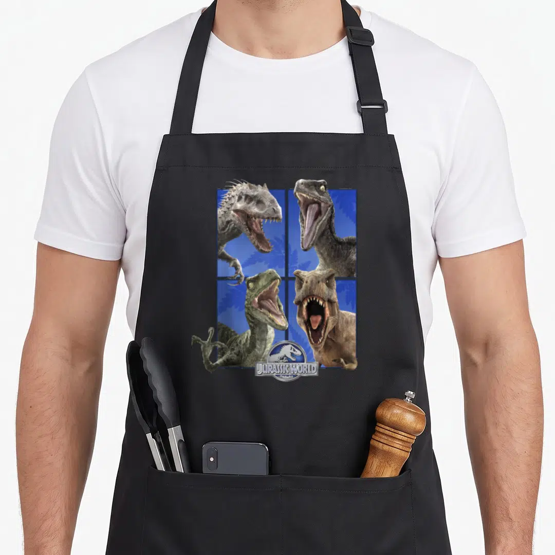 Roar dinosaur apron - Four dinosaur heads in a grid over blue background with JURASSIC WORLD logo at bottom center — shown on a black cotton canvas apron against a white background