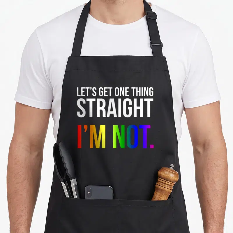 Straight rainbow apron - Stacked text reading Let's Get One Thing Straight I'm Not in white and rainbow gradient colors — shown on a black cotton canvas apron against a white background