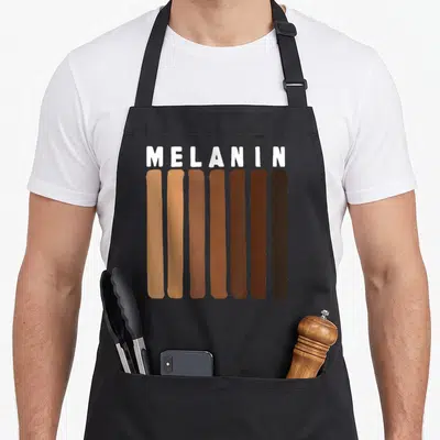 Pride melanin african print apron - MELANIN text above seven vertical skin tone swatches in tan and brown — shown on a black cotton canvas apron against a white background