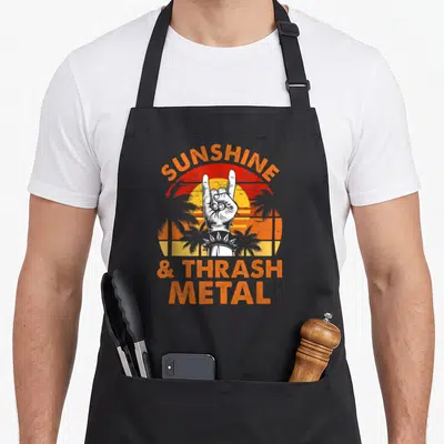 Festival heavy metal apron - Retro sun with palm trees, white rock hand sign, and orange text reading SUNSHINE & THRASH — shown on a black cotton canvas apron against a white background