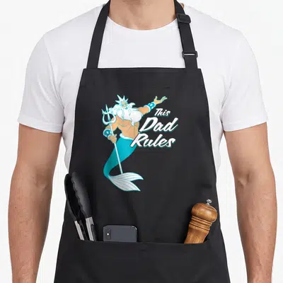 Triton king apron - Cartoon Poseidon holding a trident beside stacked white script reading This Dad Rules — shown on a black cotton canvas apron against a white background