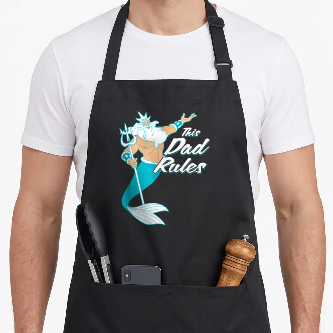 Triton king apron - Cartoon Poseidon holding a trident beside stacked white script reading This Dad Rules — shown on a black cotton canvas apron against a white background