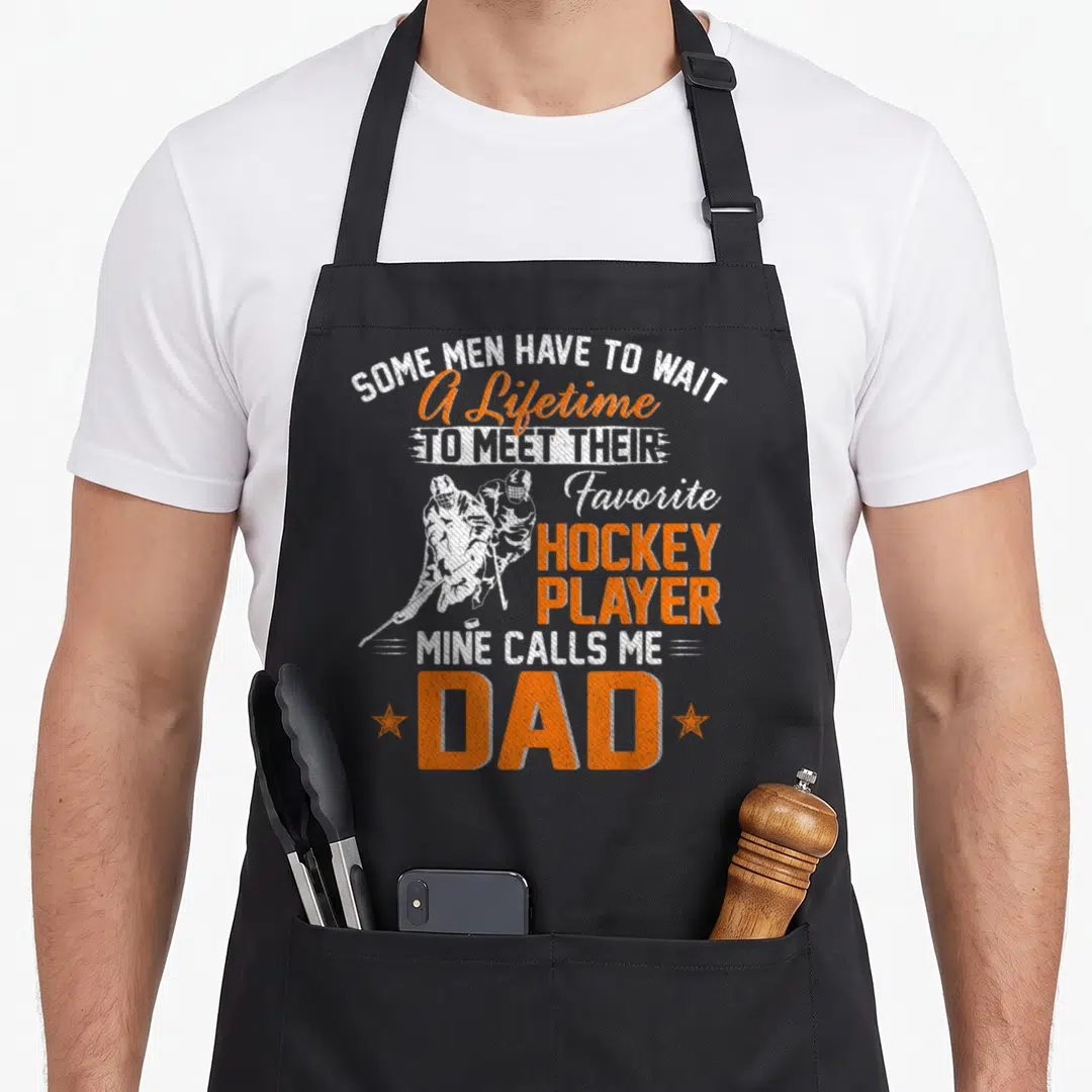 Hockey papa apron - White and orange distressed text with two hockey players and stars flanking the word Dad — shown on a black cotton canvas apron against a white background