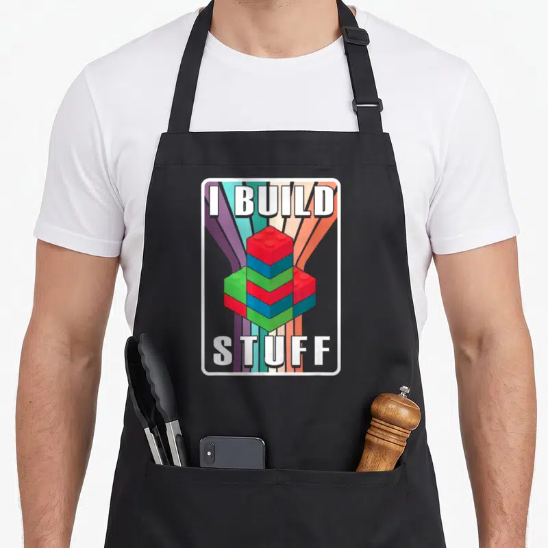 Build master block & apron - Colorful building blocks stacked between I BUILD and STUFF text over radiating stripes — shown on a black cotton canvas apron against a white background
