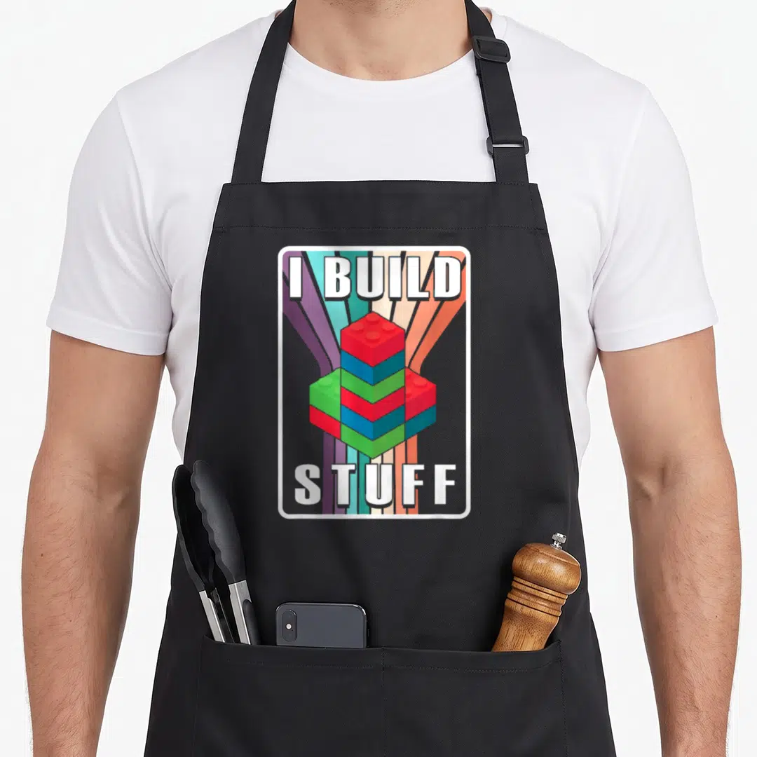 Build master block & apron - Colorful building blocks stacked between I BUILD and STUFF text over radiating stripes — shown on a black cotton canvas apron against a white background