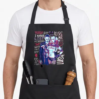 Squad vehicle apron - Joker and Harley Quinn surrounded by scattered text quotes in pink and blue distressed style — shown on a black cotton canvas apron against a white background