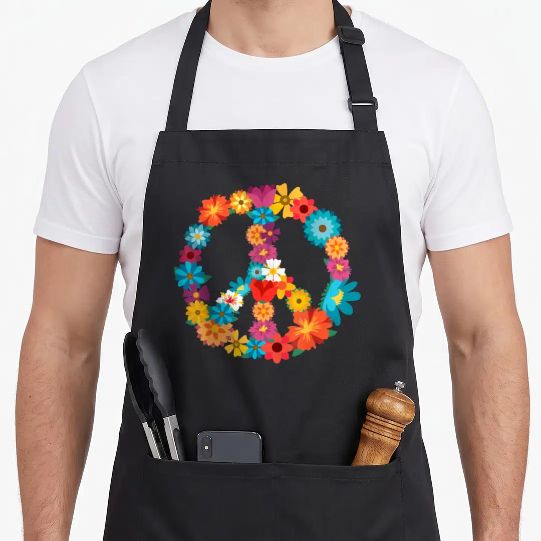 Colorful hippie apron - Peace symbol formed by red blue yellow orange pink and purple cartoon flowers — shown on a black cotton canvas apron against a white background