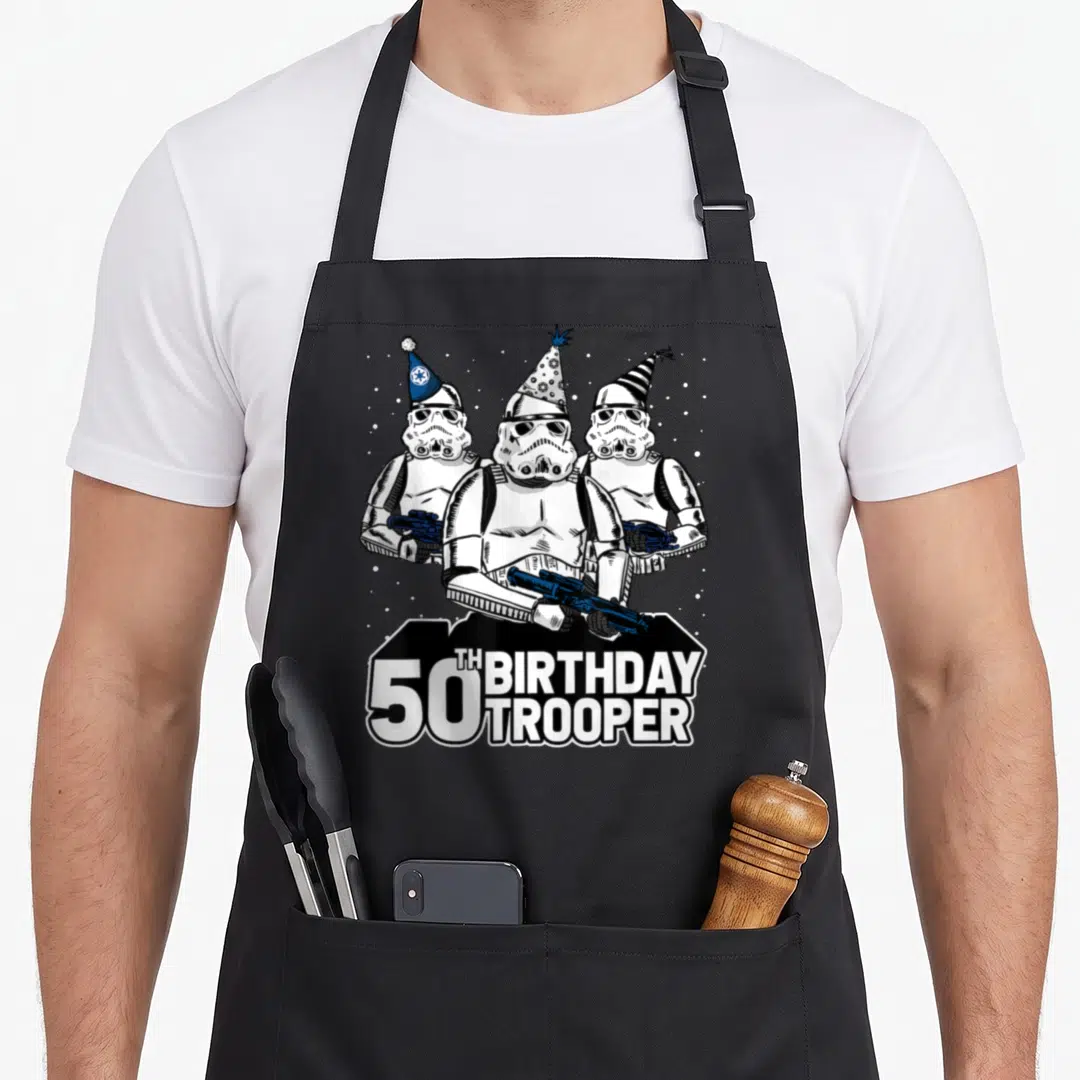 Hat birthday apron - Three stormtroopers in party hats holding blasters above 50TH BIRTHDAY TROOPER text banner — shown on a black cotton canvas apron against a white background