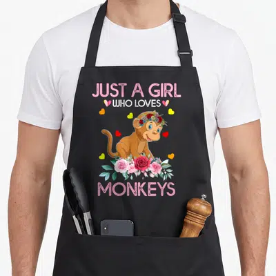 Funny monkey apron - Cartoon monkey with flower crown and roses surrounded by hearts and text Just A Girl Who Loves — shown on a black cotton canvas apron against a white background