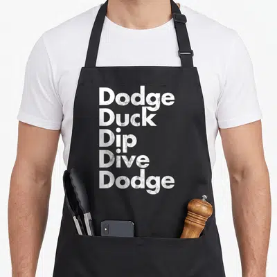 Dive duck apron - Stacked white text reading Dodge Duck Dip Dive Dodge on grey background — shown on a black cotton canvas apron against a white background
