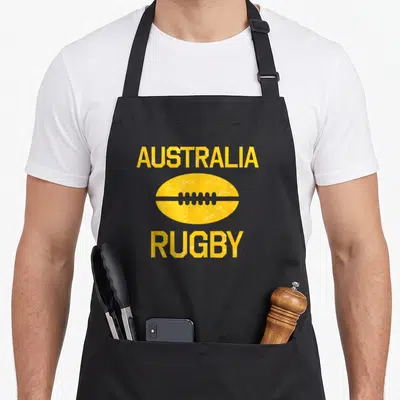 Rugby australian apron - Yellow distressed rugby ball graphic centered between AUSTRALIA and RUGBY text in block letters — shown on a black cotton canvas apron against a white background