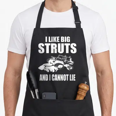 Rock truck apron - White distressed text I Like Big Struts above and below monster truck kicking up mud — shown on a black cotton canvas apron against a white background