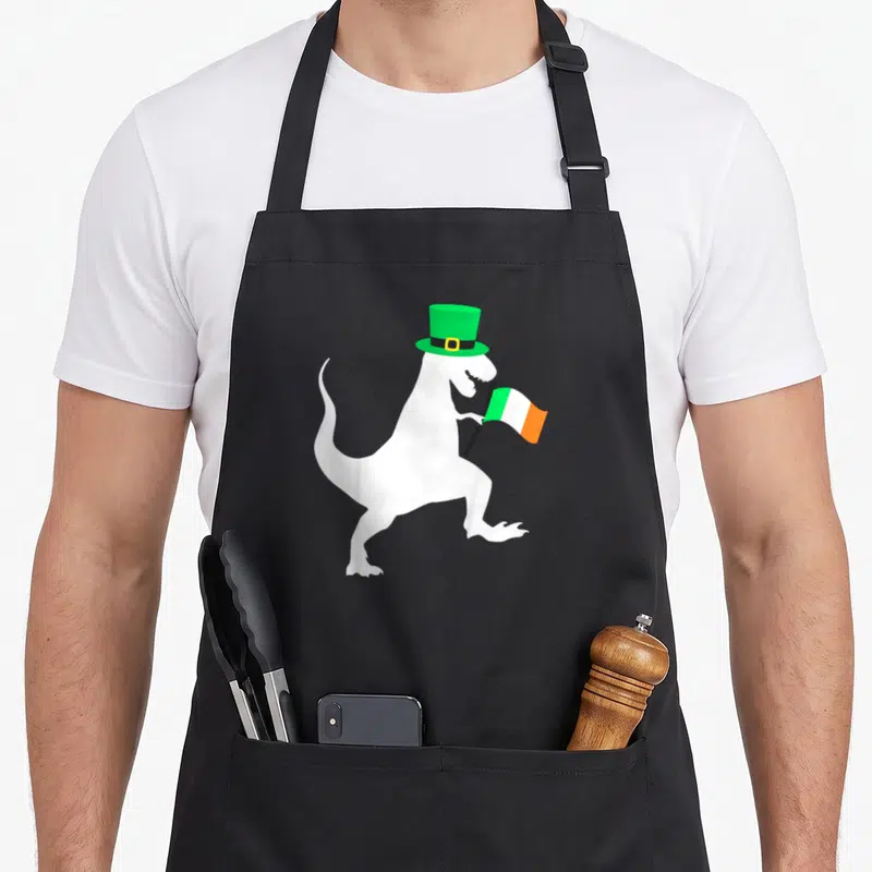 Saint dinosaur apron - White dinosaur silhouette wearing green top hat and holding green white orange flag — shown on a black cotton canvas apron against a white background