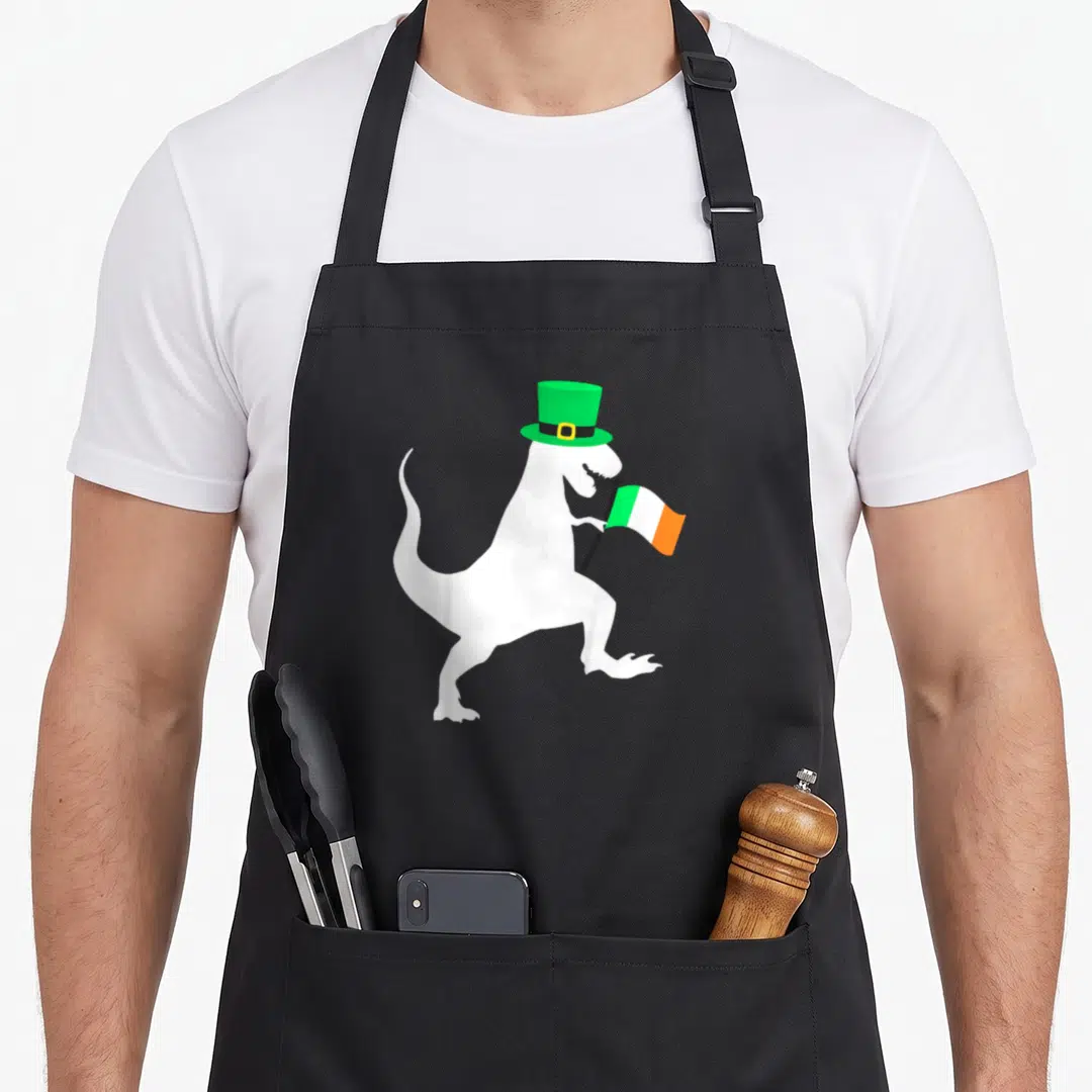 Saint dinosaur apron - White dinosaur silhouette wearing green top hat and holding green white orange flag — shown on a black cotton canvas apron against a white background