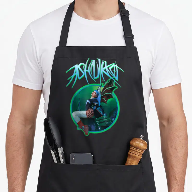 Ashnikko dragon apron - Arched ASHNIKKO text above winged woman in glowing circle with blue, green, red, and black — shown on a black cotton canvas apron against a white background