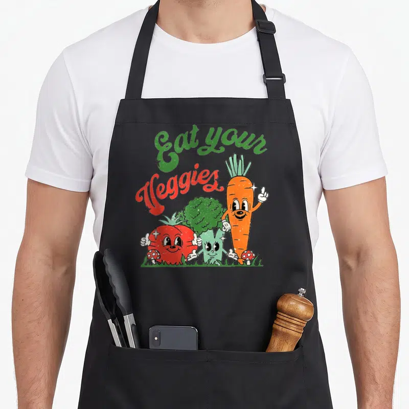 Eat vegetable apron - Cartoon carrot tomato broccoli with Eat your Veggies text and mushrooms on grass — shown on a black cotton canvas apron against a white background