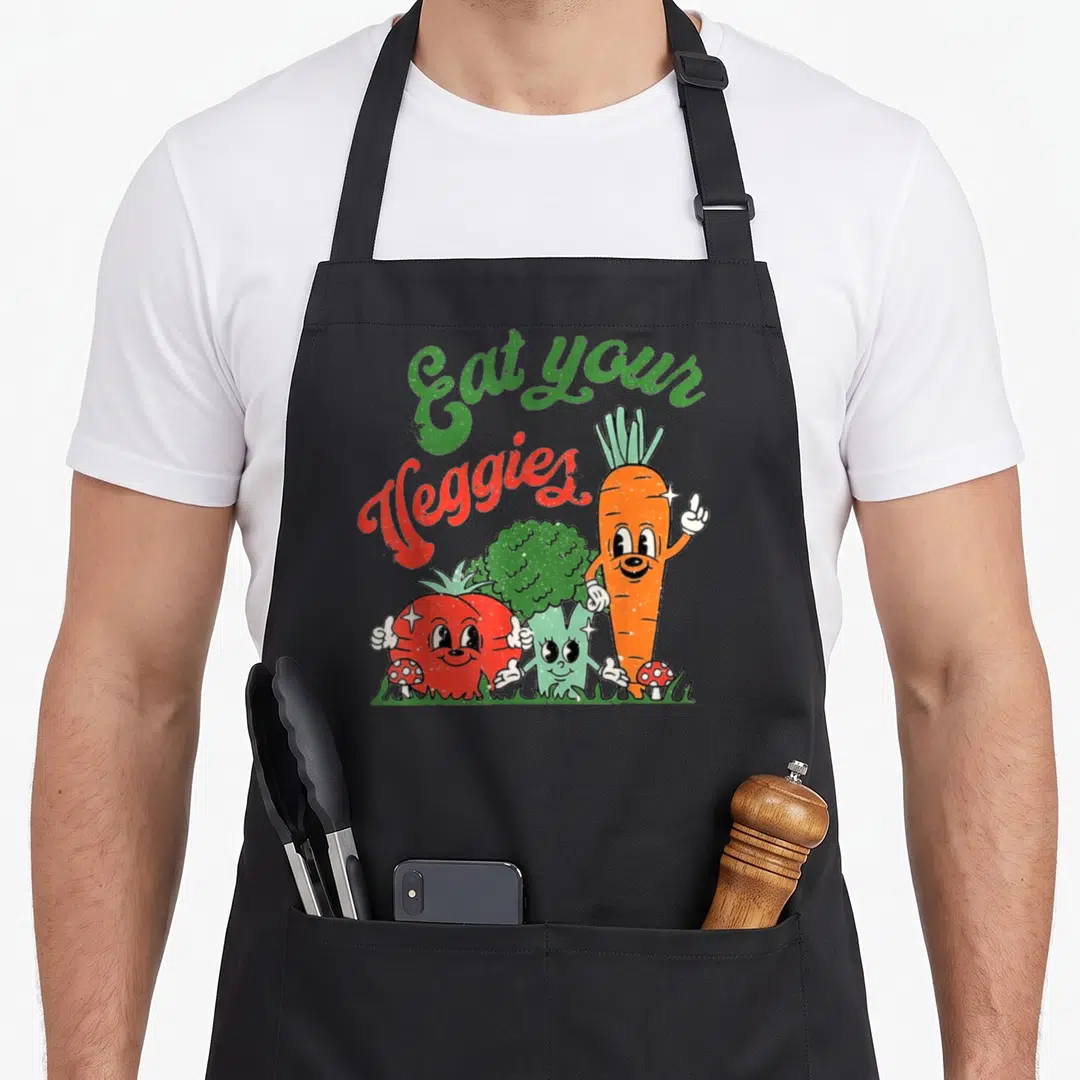 Eat vegetable apron - Cartoon carrot tomato broccoli with Eat your Veggies text and mushrooms on grass — shown on a black cotton canvas apron against a white background