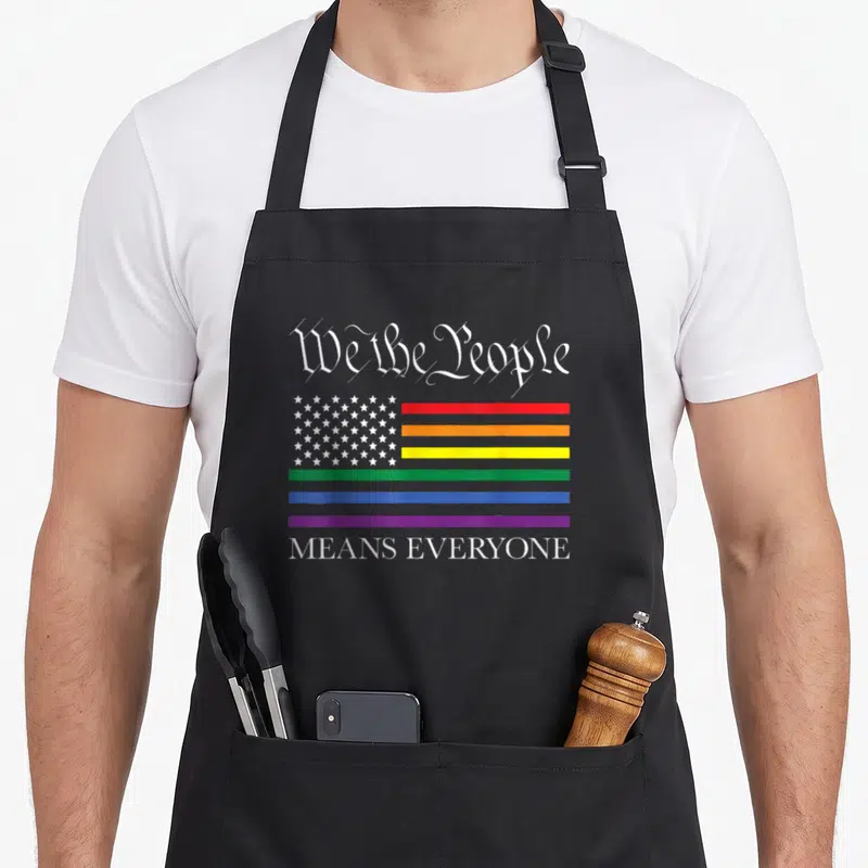 Equality gay pride apron - We the People text above rainbow flag with white stars and Means Everyone slogan below — shown on a black cotton canvas apron against a white background