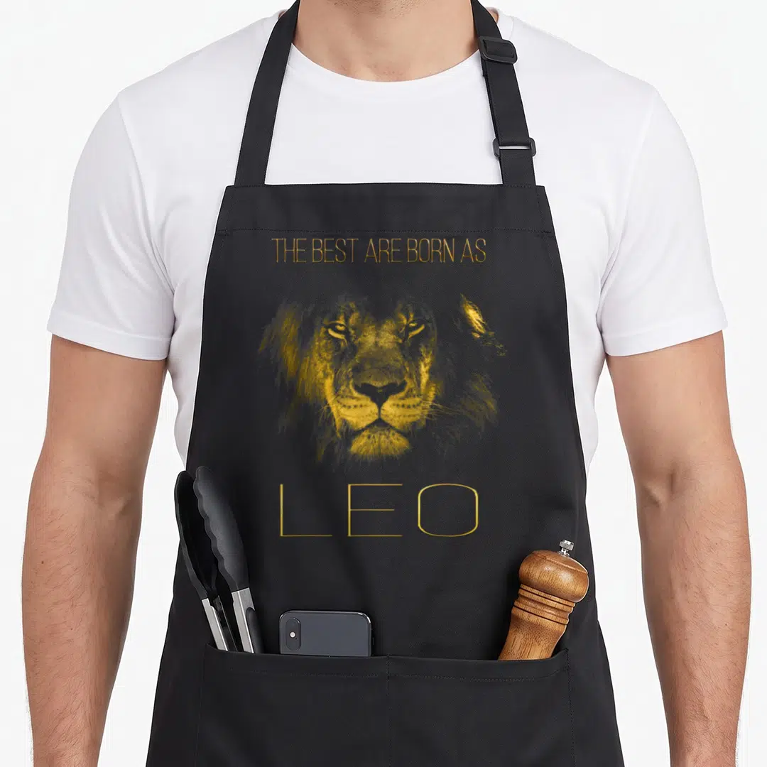 Leo lion apron - Realistic gold lion face with text The Best Are Born As Leo above and below — shown on a black cotton canvas apron against a white background