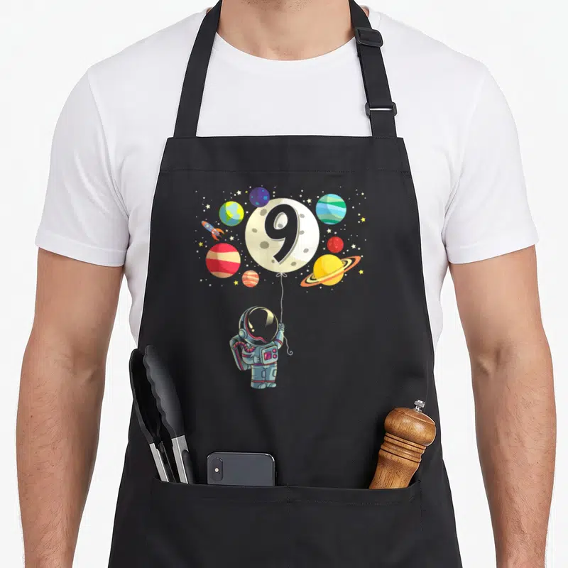 Astronaut 9th birthday apron - Cartoon astronaut holding a moon balloon with the number 9 surrounded by planets — shown on a black cotton canvas apron against a white background