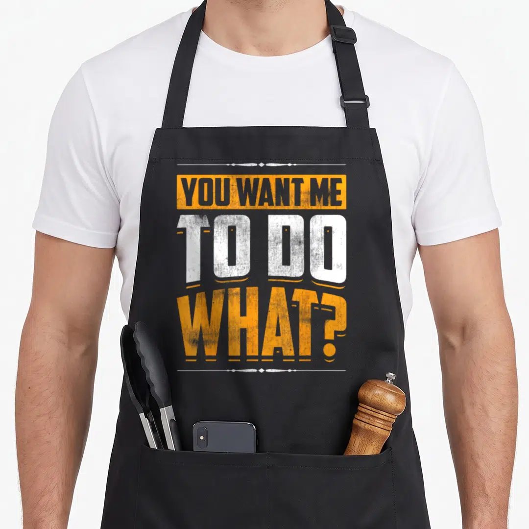 Associate warehouse problems apron - Stacked distressed text YOU WANT ME TO DO WHAT in orange and white blocks — shown on a black cotton canvas apron against a white background