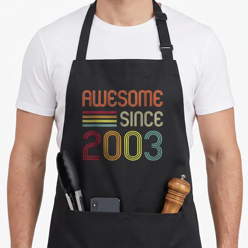 Funny 20th birthday apron - Retro AWESOME SINCE 2003 text with orange beige red yellow teal horizontal lines — shown on a black cotton canvas apron against a white background